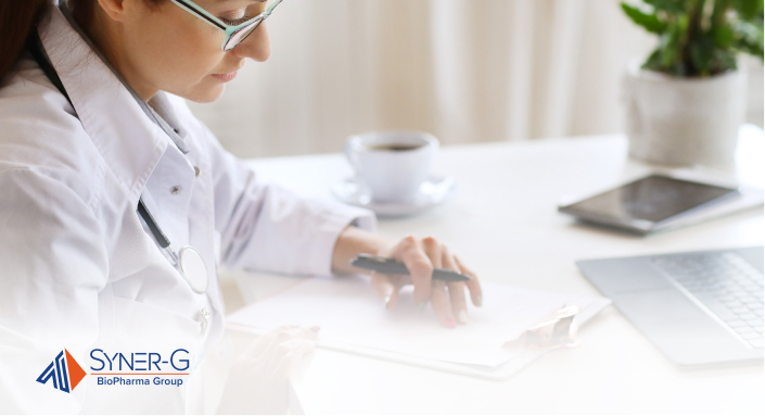 A female doctor in a white coat writes notes at a desk with a stethoscope and computer, set in a bright clinic office.